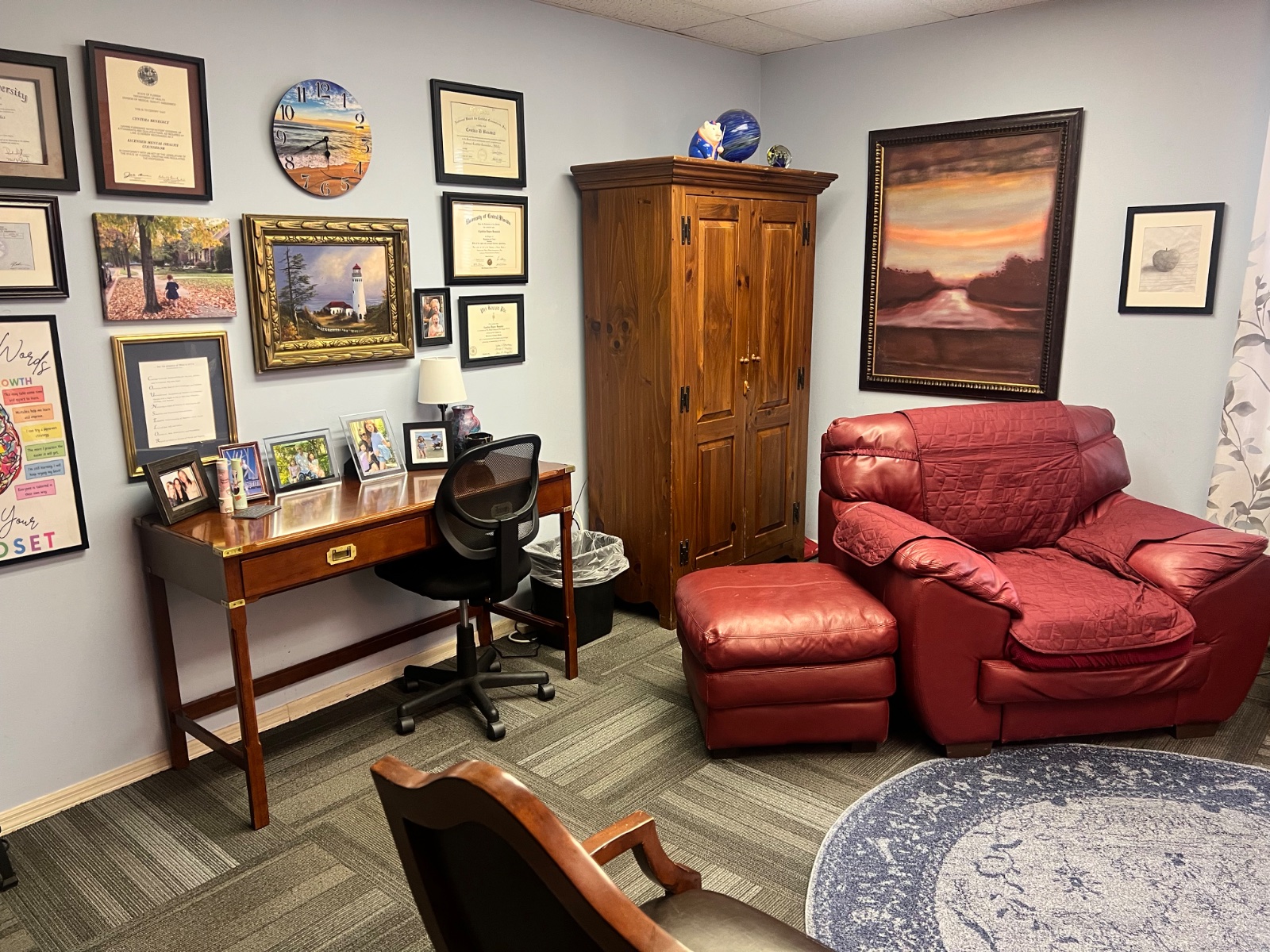 Cozy seating area in Cynthia Benedict's Longwood counseling office with a red leather chair, wooden armoire, and framed artwork